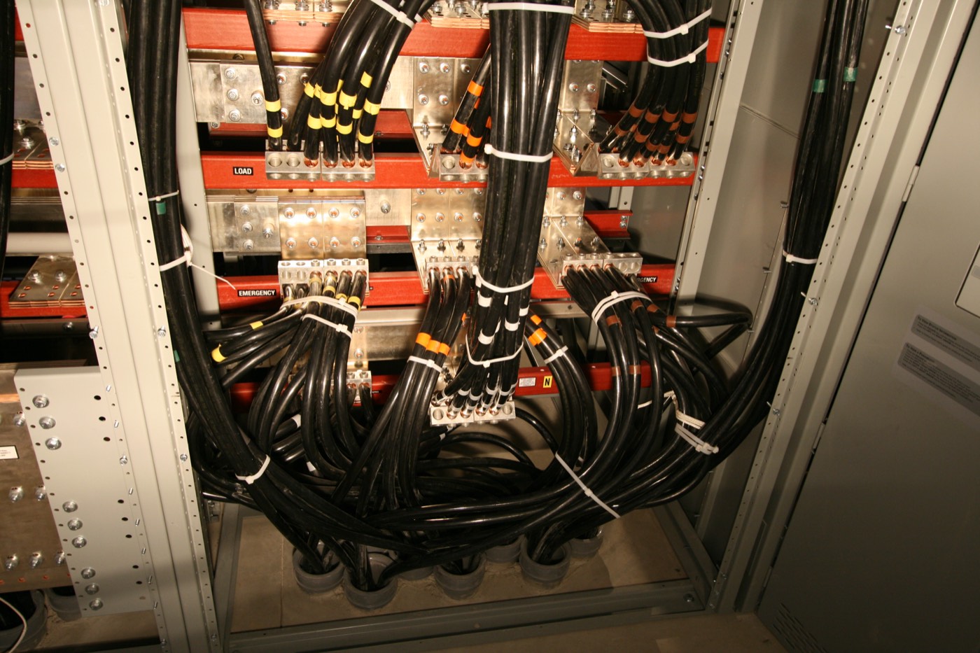 Close-up of switchgear terminations: copper busbar with load and emergency labels and lugged feeder cables.