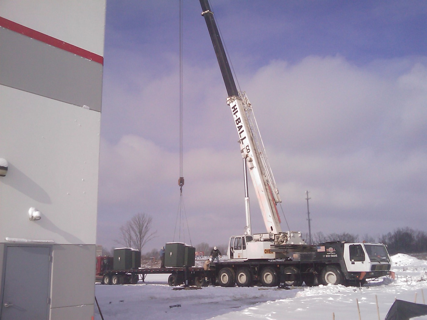 Mobile crane offloading electrical equipment from flatbed trailers outside the building in winter.