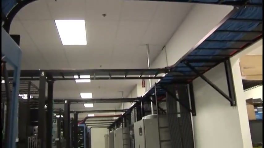 A data hall aisle with equipment cabinets along the wall and overhead cable tray carrying bundled network cabling.