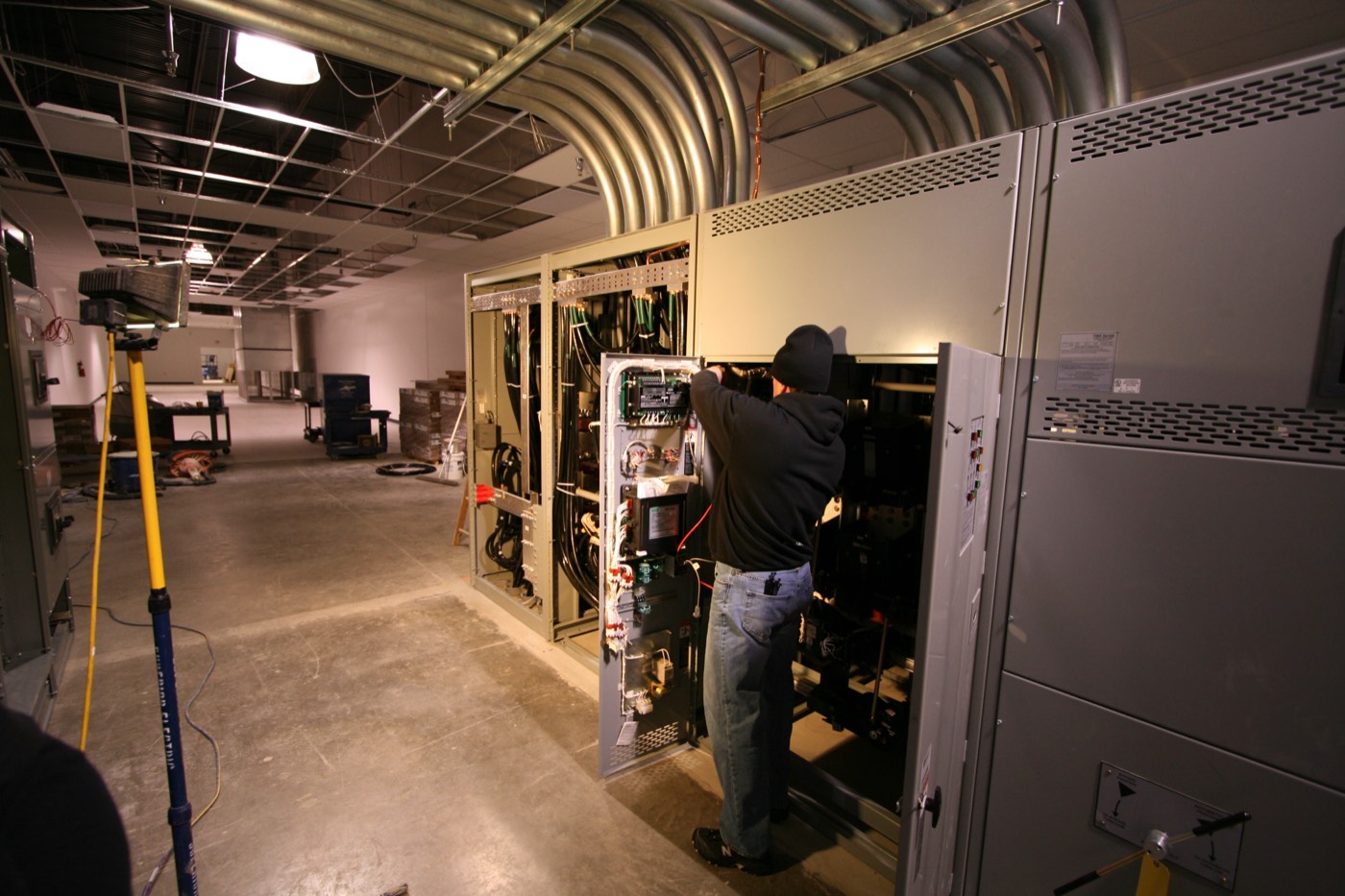 Electrician working inside an open switchgear section, banks of conduit entering the electrical room overhead.